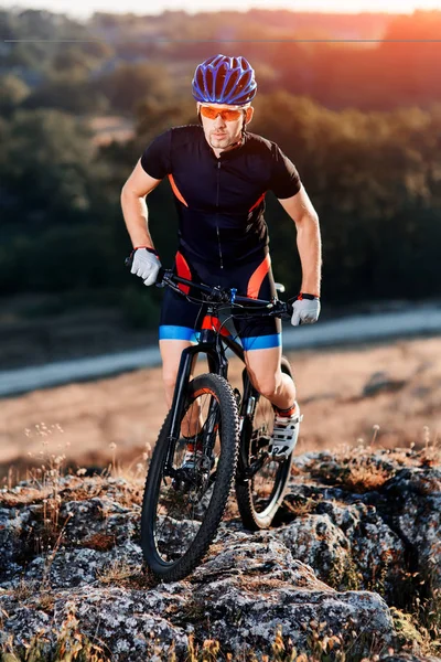 Ciclista profesional en casco azul y gafas de sol naranjas Montar en bicicleta en la cima de la roca. Concepto de deporte extremo . — Foto de Stock