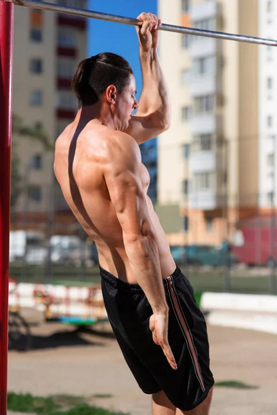 Giovane atleta muscolare che fa esercizi di pull-up appeso con una mano dritta su una barra orizzontale. Foto dal retro . — Foto Stock