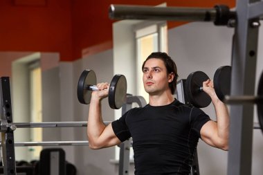 Genç kas atlet bir halter makine üzerinde oturan ve iki halter kaldırma siyah eşofman içinde inşa