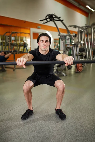 Young athletic man in black sportwear with bar of barbell flexing muscles in gym.