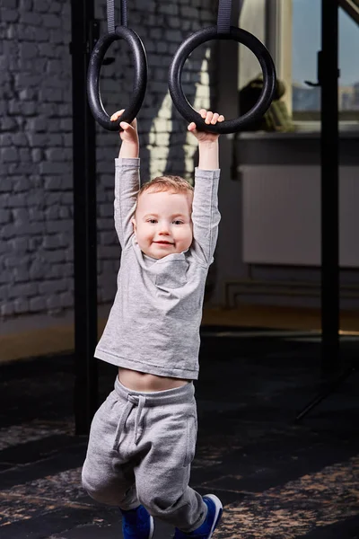 Pequeño chico deportivo en la ropa deportiva gris colgando de anillos de gimnasia en el gimnasio . — Foto de Stock