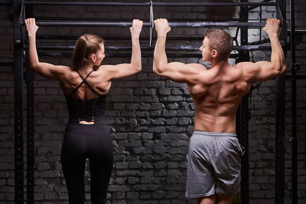 Retrovisore foto di muscoloso uomo e donna che fa esercizi su barra orizzontale contro muro di mattoni alla palestra cross fit . — Foto Stock