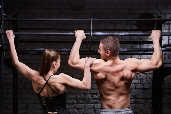Retrovisore foto di muscoloso uomo e donna che fa esercizi su barra orizzontale contro muro di mattoni alla palestra cross fit . — Foto Stock