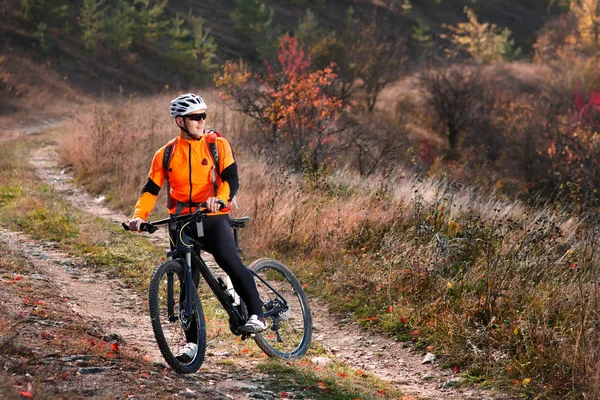 Ciclista en la chaqueta naranja montando una bicicleta en la carretera rural al amanecer . — Foto de Stock