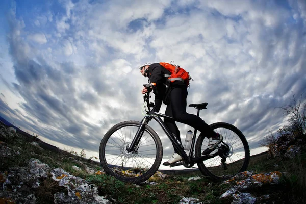 Ciclist în îmbrăcăminte sport negru de echitatie bicicleta pe stâncă seara împotriva cer albastru frumos cu nori . — Fotografie, imagine de stoc