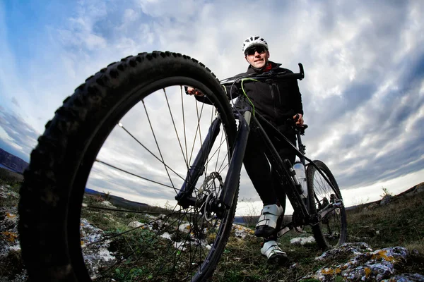 Фотографія велосипедиста в чорному спортивному одязі, що стоїть зі своїм велосипедом на скелі проти блакитного неба з хмарами . — стокове фото
