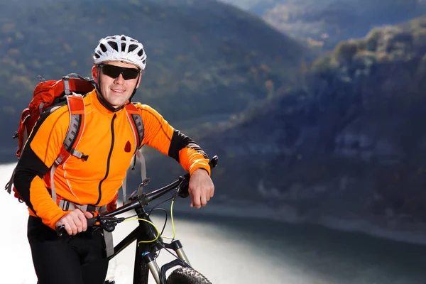 Mountain cyclist in the orange jacket stands under the beautiful river against mountains.