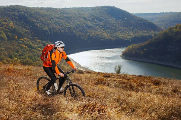 Cyclist in orange sporyive jacket riding the bike on the hill under river against beautiful mountain.