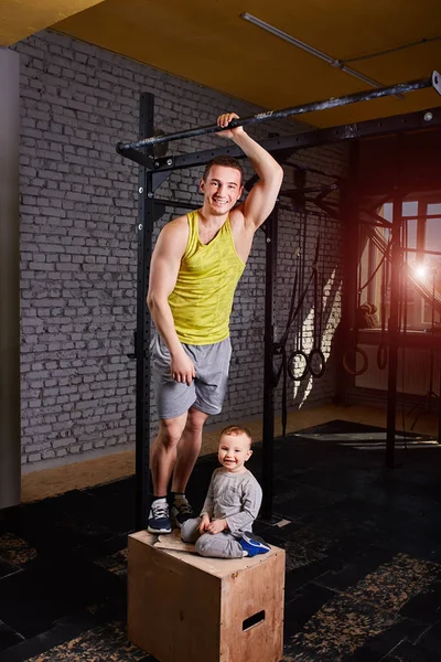 Lächelnder junger Vater mit kleinem Sohn steht auf der Kiste an der Ziegelwand der Crossfit-Turnhalle. — Stockfoto