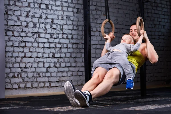 Genç atlet baba ve gimnastic halkalar ile egzersiz ve tuğla karşı gülümseyen küçük sevimli oğlu salonda duvar.