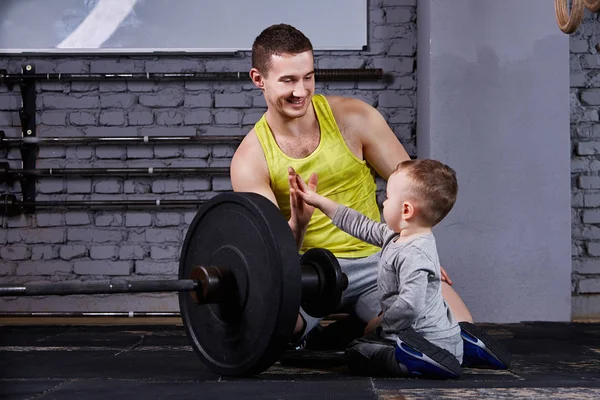 Jovem pai desportivo e pequeno filho bonito sentado perto barbell contra parede de tijolo na cruz caber ginásio . — Fotografia de Stock