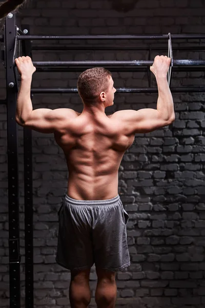 Rear view photo of the young muscular male doing exercises on horizontal bar against brick wall at the cross fit gym.