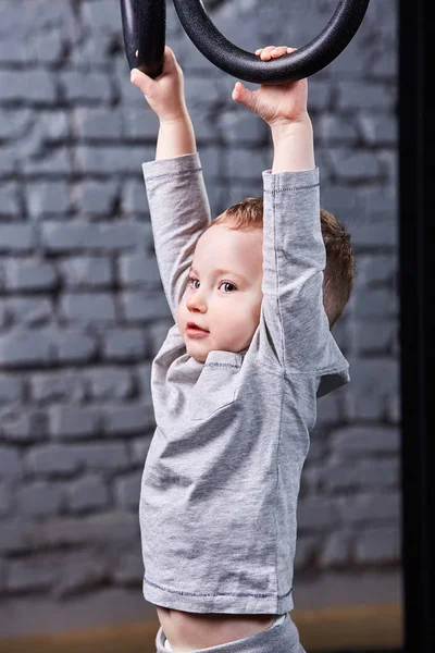 Niño en ropa deportiva gris tge colgando de anillos de gimnasia contra la pared de ladrillo en el gimnasio — Foto de Stock