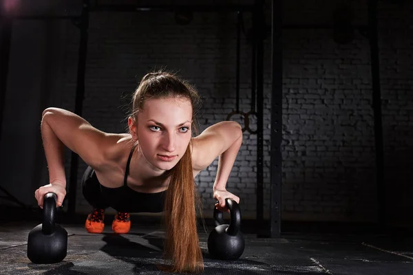 Young sporty woman doing push-ups exercise with kettlebell against brick wall.