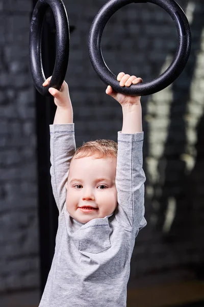 Lindo niño colgando de anillos de gimnasia en el gimnasio cross fit contra la pared de ladrillo . — Foto de Stock