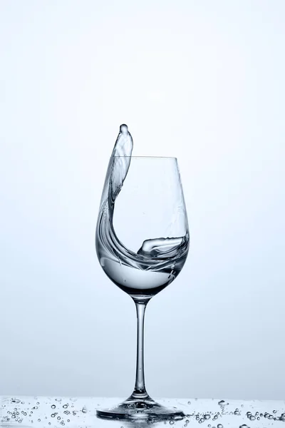 Vatten stänk från vinglas med renare vatten stående på glaset med droppar mot ljus bakgrund. — Stockfoto
