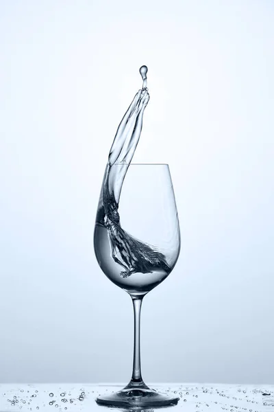Cleaner water splash out of wineglass while standing on the glass with droplets against light background.