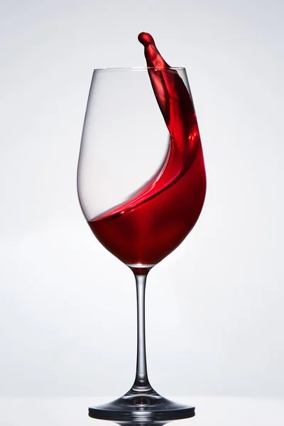 Červené víno stříkající z čisté elegantní skleničku stojící proti světlé pozadí s odleskem. — Stock fotografie