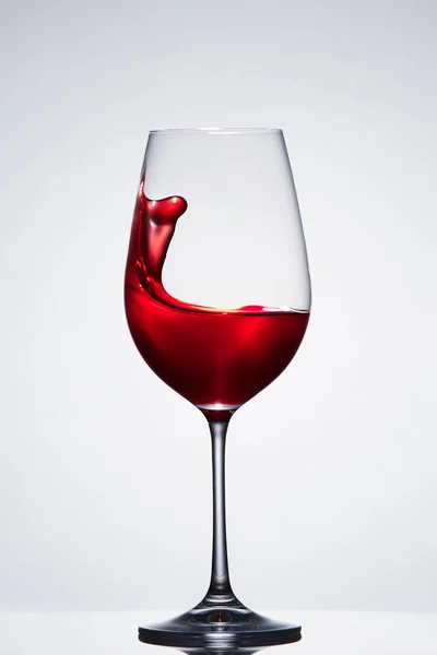 Červené víno stříkající z čisté elegantní skleničku stojící proti světlé pozadí s odleskem. — Stock fotografie