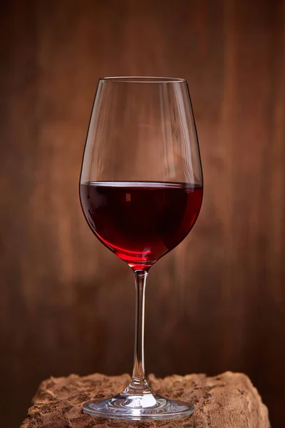 Close-up of crystal half-ful wineglass with red wine standing on wooden stand against wooden background.