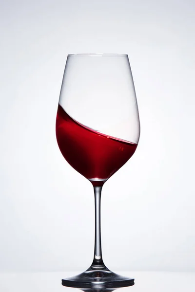 Moving red wine in the elegant wineglass standing against light background with reflection. — Stock Photo, Image