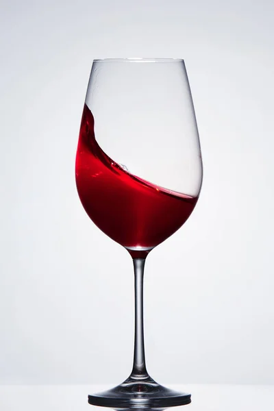 Splashing red wine with wave in the elegant wineglass standing against light background with reflection. — Stock Photo, Image
