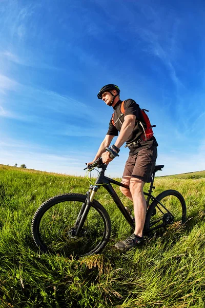 Νέος ποδηλάτης με το κράνος στο πράσινο λιβάδι σε φόντο με μπλε του ουρανού. — Φωτογραφία Αρχείου