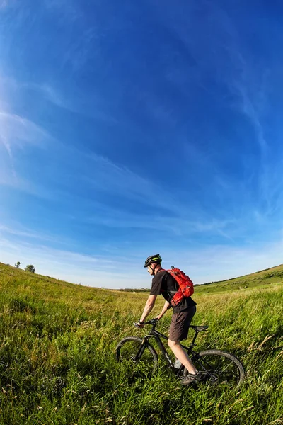 Mladý cyklista v helmě na zelené louce na pozadí s modrou oblohou. — Stock fotografie