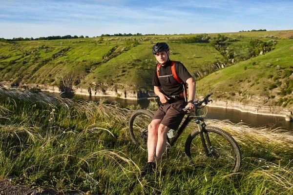 Retrato del joven ciclista de pie en la colina con bicicleta sobre el río . — Foto de Stock