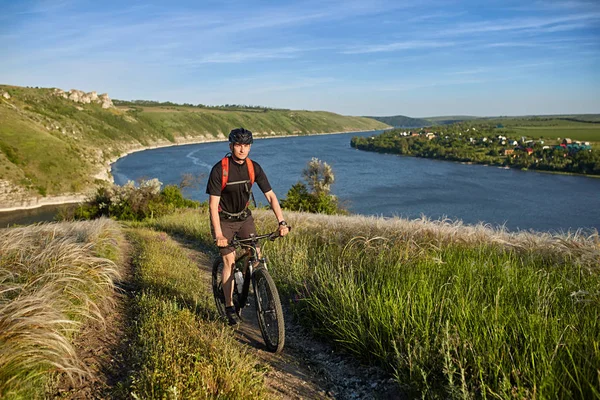 Mladý cyklista jízdní horské kolo do kopce podél venkovské silnici nad řekou. — Stock fotografie