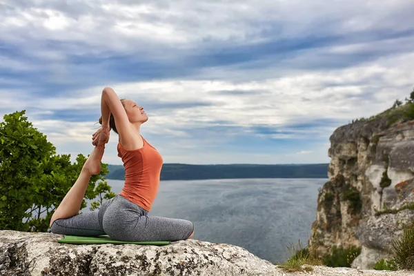 Молодая женщина делает йогу упражнения голубя asana на скале против реки . — стоковое фото