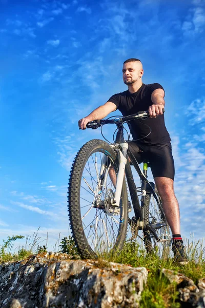 Vista inferior do jovem ciclista de pé com bicicleta em rochas contra o céu azul . — Fotografia de Stock