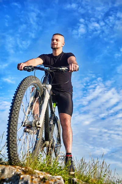 Vista inferior do jovem ciclista de pé com bicicleta em rochas contra o céu azul . — Fotografia de Stock