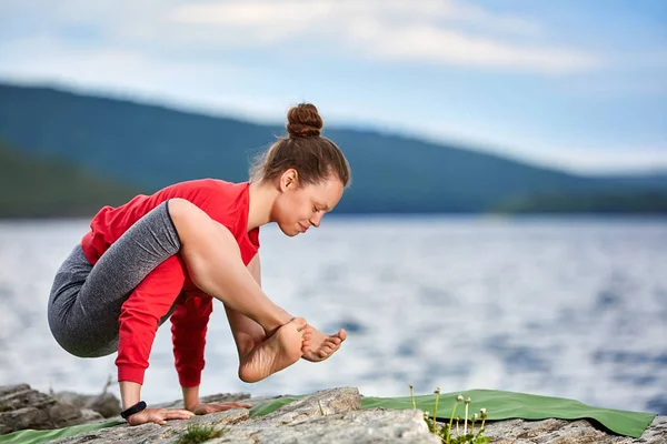 Молодая женщина делает упражнения йоги на камне возле большой реки против красивого ландшафта . — стоковое фото