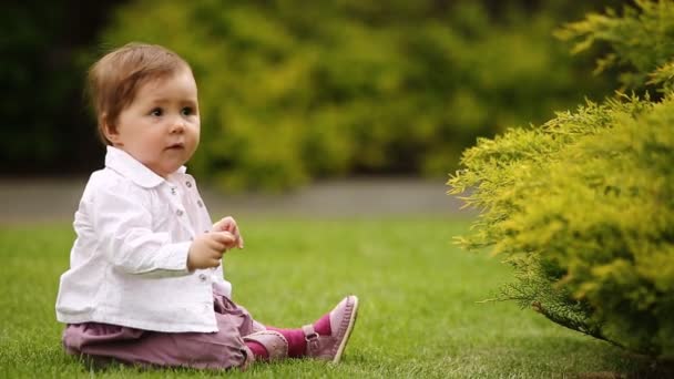 Веселая девочка сидит на зеленой траве рядом с кустом в городском парке . — стоковое видео
