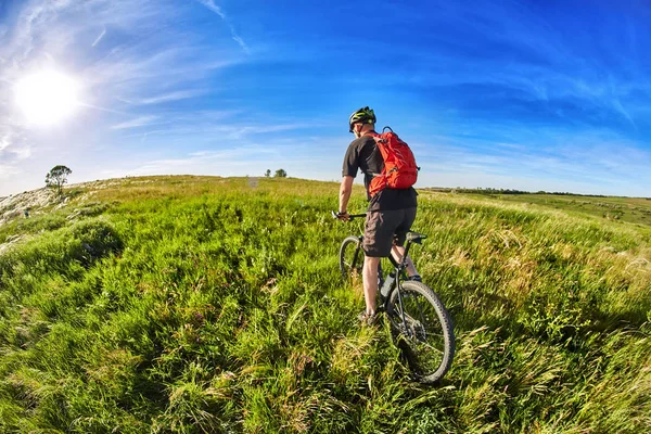 Νέος ποδηλάτης ιππασίας ποδήλατο βουνού μέσω πράσινο λιβάδι ενάντια όμορφο ουρανό. — Φωτογραφία Αρχείου