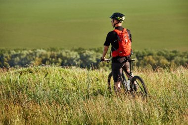 Arkadan görünüş onun dağ bisikleti ile sporcu, çayır ve uzak kırsal kesimde seyir üzerinde duruyor.