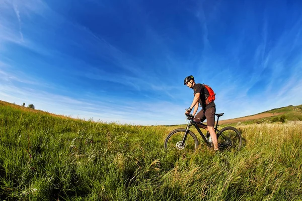 Giovane ciclista in bicicletta sul prato verde contro il bel cielo blu con nuvole in campagna . — Foto Stock