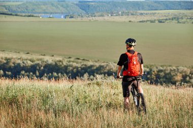 Arkadan görünüş onun dağ bisikleti ile sporcu, çayır ve uzak kırsal kesimde seyir üzerinde duruyor.