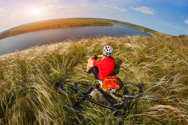 Der attraktive Radfahrer sitzt auf dem Gras auf der Wiese mit dem Mountainbike über dem Fluss. — Stockfoto