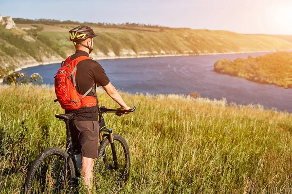 Pohled zezadu na mladý cyklista porostů s horské kolo na zelené louce nad velké řeky. — Stock fotografie