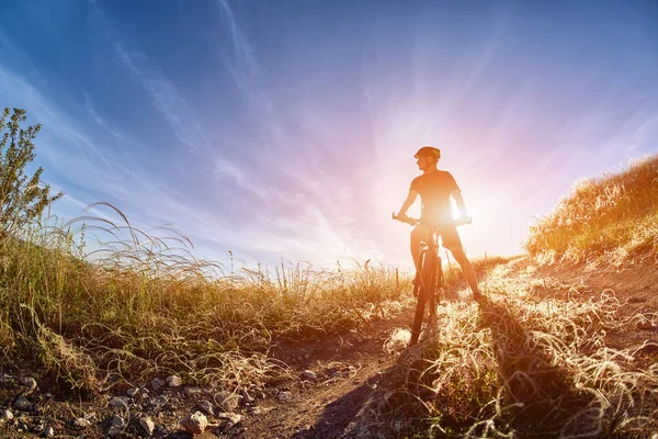 Jovem ciclista com ciclista de montanha no caminho do campo no campo contra o nascer do sol . — Fotografia de Stock