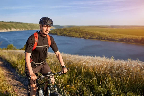 Retrato del joven ciclista de pie en la colina sobre el río contra el cielo azul con nubes . — Foto de Stock