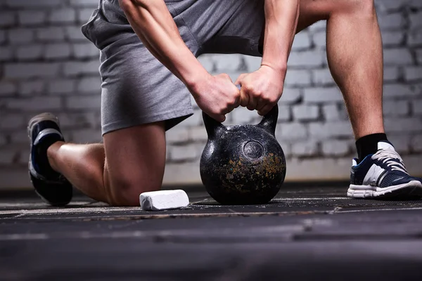 Close-up photo of young sportsman while crouching on a one leg and holding kettlebell against brick wall. — Stock Photo, Image