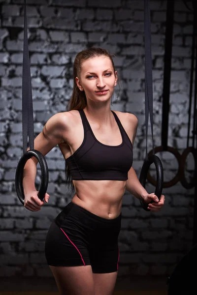Image of beautiful young sporty woman in the black sportwear doing pull-ups exercise using gymnastic rings.