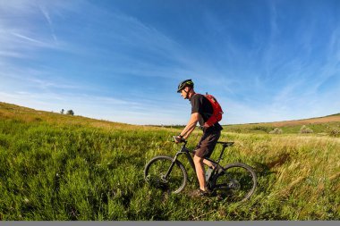 Genç adam bir sırt çantası ile yeşil çayır bir bisiklet sürme