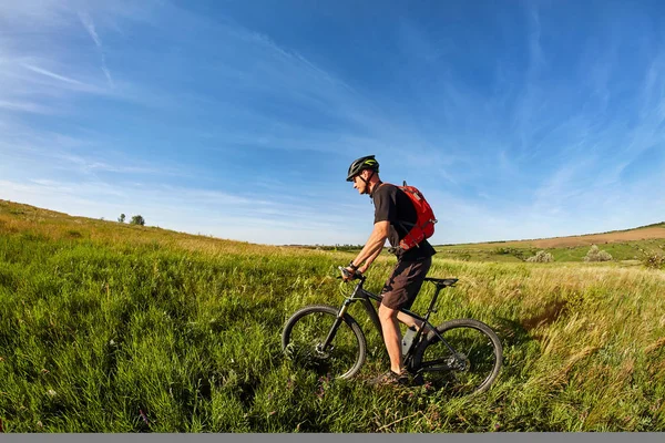 Joven montado en una bicicleta en el prado verde con una mochila — Foto de Stock