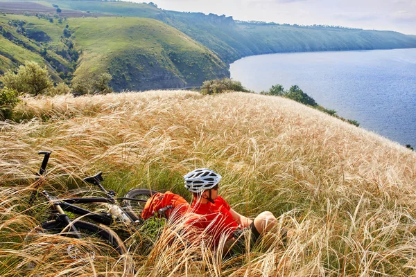Atraktivní cyklista sedí na trávu na louce s horské kolo nad řekou. — Stock fotografie