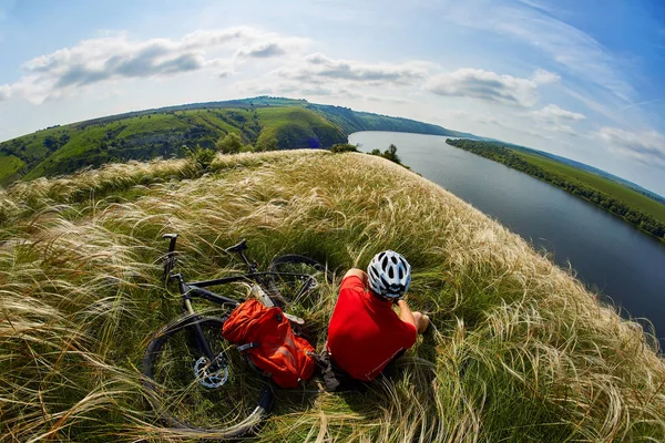 Der attraktive Radfahrer sitzt auf dem Gras auf der Wiese mit dem Mountainbike über dem Fluss. — Stockfoto