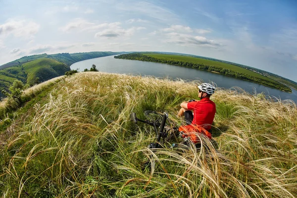 Der attraktive Radfahrer sitzt auf dem Gras auf der Wiese mit dem Mountainbike über dem Fluss. — Stockfoto